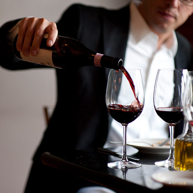 A man in a suit pours red wine into two glasses on a table.