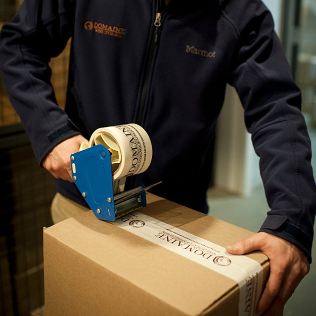 A man seals a box of wine using Domaine tape, packing it before our wine transit team delivers it.