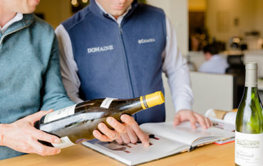 Two Domaine employees perform wine appraisals by examining a wine bottle and catalogue.