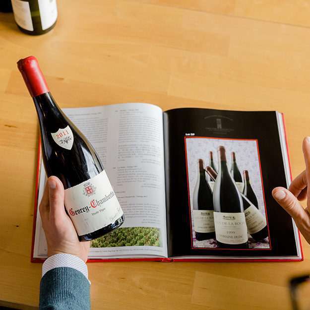 A man holds a bottle of red wine next to an open magazine with the same bottle advertised for those who enjoy buying wine on its pages.