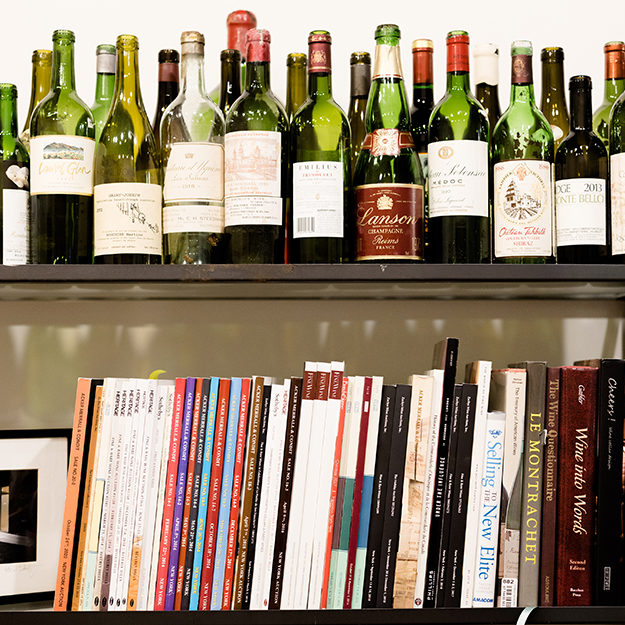 A row of books about wine and wine cellar management on a shelf, with wine bottles on the shelf above.
