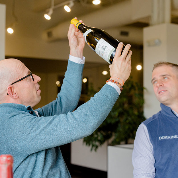 Two of Domaine's wine selling experts hold up and inspect a bottle of wine in the light.